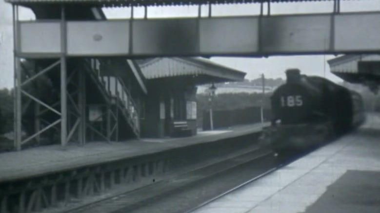 Black and white image of an old steam train pulling up to a platform.