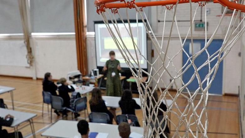 A picture of a basketball hoop in Myton School's sports hall. In the background, out of focus, a group of about a dozen students are learning at foldable desks.