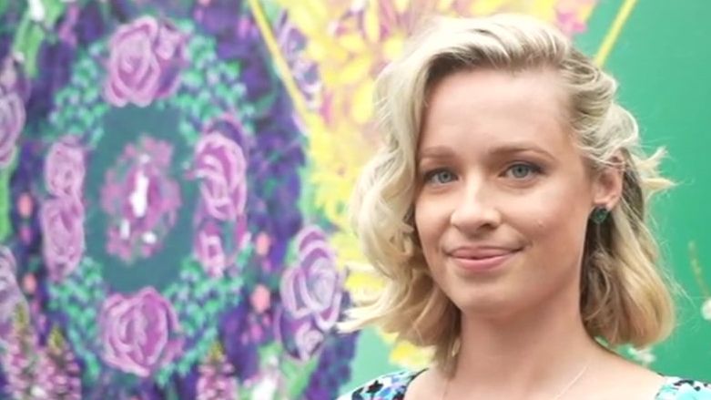 A head-and-shoulders image of a woman with short blond hair. She is looking at the camera and smiling. Behind her is a mural, which is blue, yellow and green.