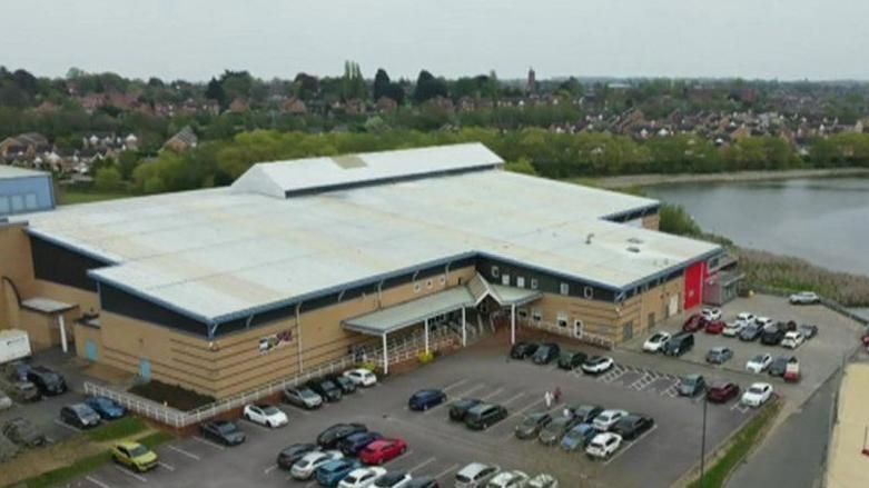 Large brown building with white roof and covered entrance. There is a large car park in front of it and another building to the right.