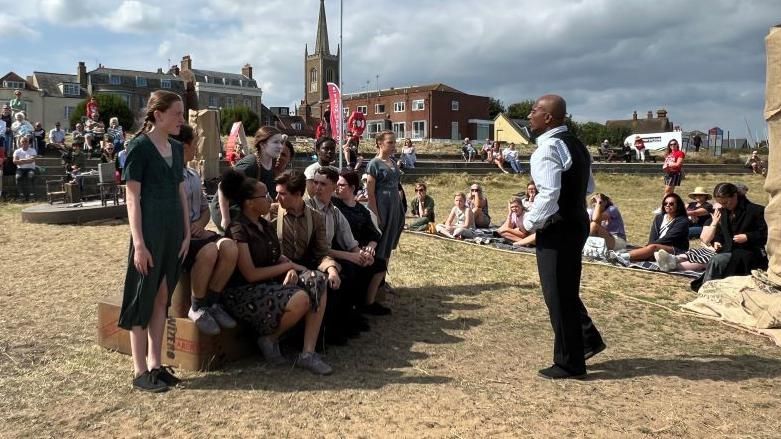 Children taking part in an outdoor performance. They are wearing 1930s-style clothes and are watched by a small audience