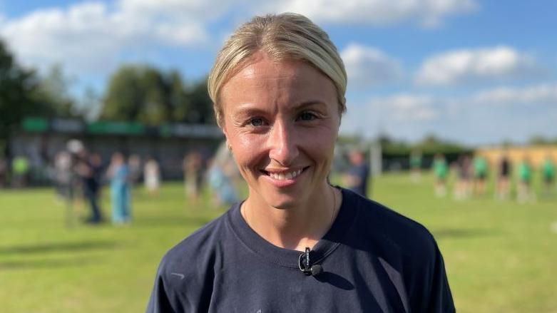 A smiling Leah Williamson looking directly at the camera with a football pitch behind her.