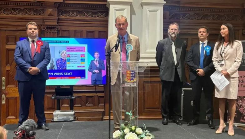 Rupert Lowe at a podium after winning the seat of Great Yarmouth on 4 July. he is wearing a brown suit and a pink tie and dons a Reform UK rosette