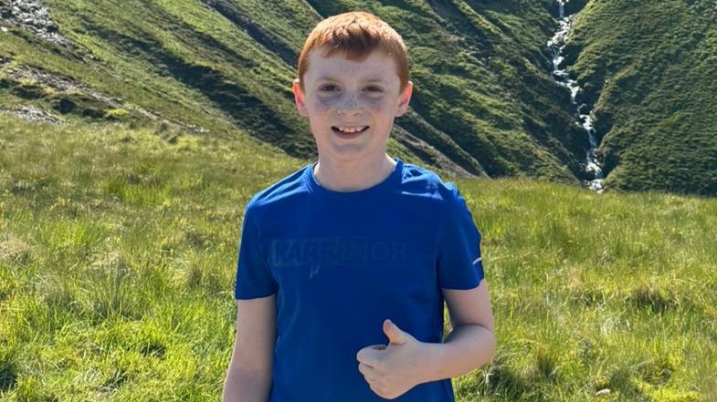 Alex standing in front of Ben Nevis. He had red hair and is wearing a blue T-shirt while giving a thumbs up to the camera. Behind him on the mountain, a stream runs along a valley down the grassy side of the mountain