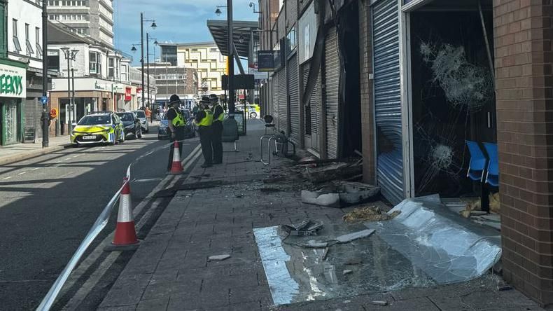 Broken window on the ground in front of a police office. Police officers are standing further up the street and debris from the burned out unit next door spills out on to the pavement.