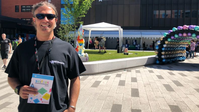 Dr Martin Khechara smiles while holding a booklet about the festival in front of a gazebo and inflatable balloon entrance way. He is wearing a black T-shirt and sunglasses