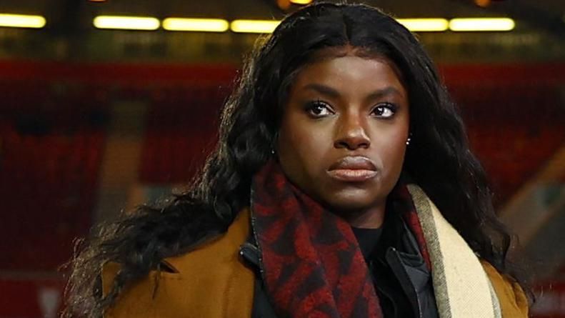 Eni Aluko, with long black hair and wearing a red and black scarf, brown overcoat and black jacket, looks up and over the camera in a football stadium