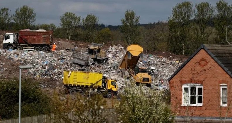 Four dumper trucks depositing waste material on a mound with trees and a red brick building in the foreground