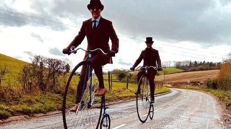 Two men in tweed suits and top hats riding penny farthings down a country road