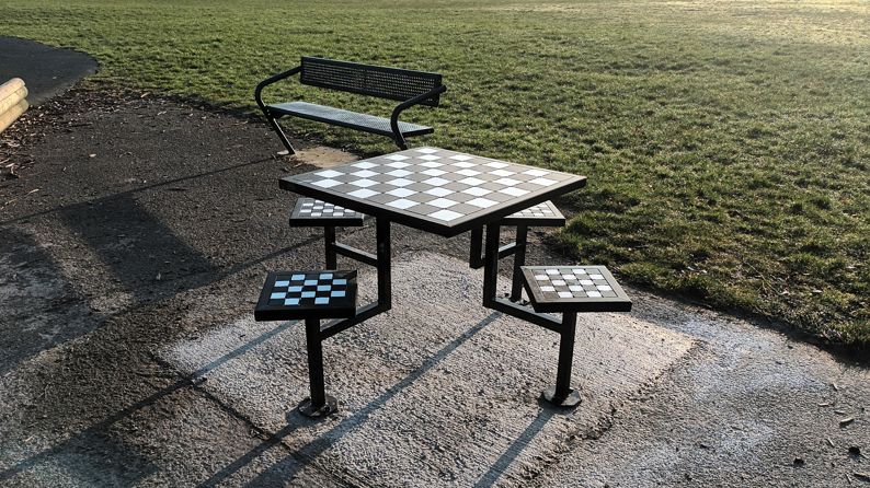 The four-seater metal table, which feature a chess board on top, also has a chess board design on each seat. It's on a gravel area, surrounded by a playing field.