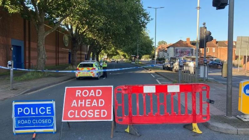 A blocked off road with police tape, a barrier, a red Road Ahead Closed sign and a blue Police Road Closed sign. There is a police car and officer in the background