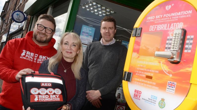 Sergio Petrucci from the Red Sky Foundation, Kelly Chequer, Chair of Sunderland City Council's Health and Wellbeing Board and Copt Hill ward councillor Kevin Johnston standing next to defibrillator