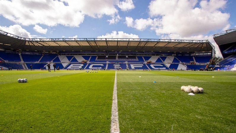 The Birmingham City football pitch from ground level, with the blue stadium seats rising from the back of the pitch saying 'BCFC'