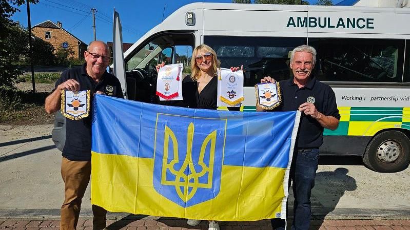 David and Terry holding a blue and yellow banner with a woman in front of the ambulance