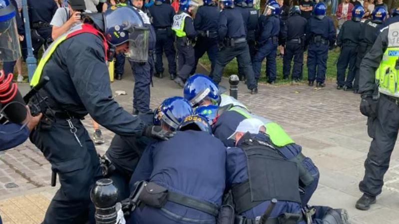 A man lying on the ground with police wearing riot gear kneeling on top of him. Officers in the background keep people at bay.