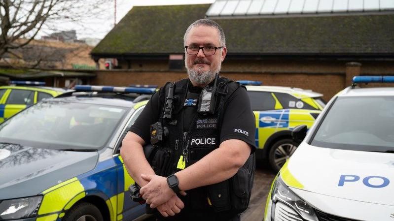 PC O'Brien with short grey hair and beard alongside police cars