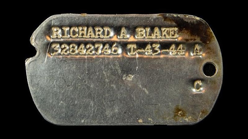 A small metal plate engraved with a name and number in the shape of a dog tag
