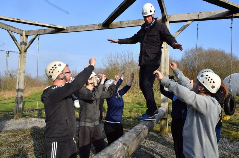 Prince William balances on a beam, supported by children, as part of an obstacle course