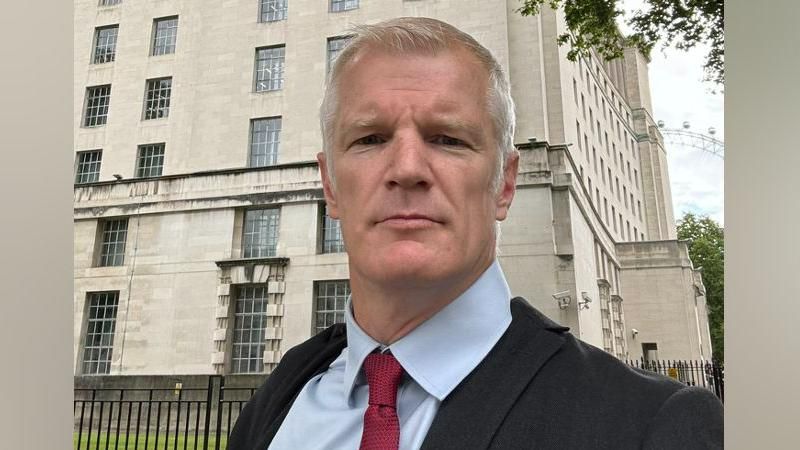 A selfie of Alistair Carns, he is wearing a grey suit jacket, light blue shirt and red tie and is standing in front of a building