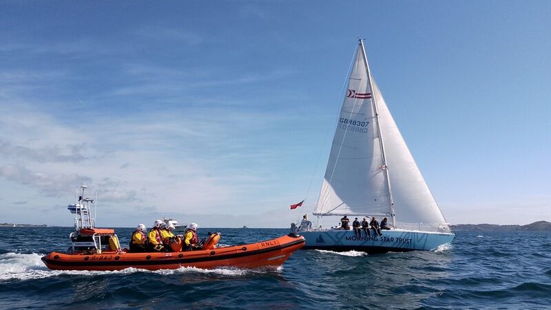 A small sailing ship sails past the Channel Island of Herm, flanked by an inflatable inshore lifeboat.