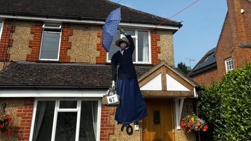 A Mary Poppins scarecrow, flying over a house, wearing a blue outfit and holding a blue umbrella, with a bag, with 67 written on it