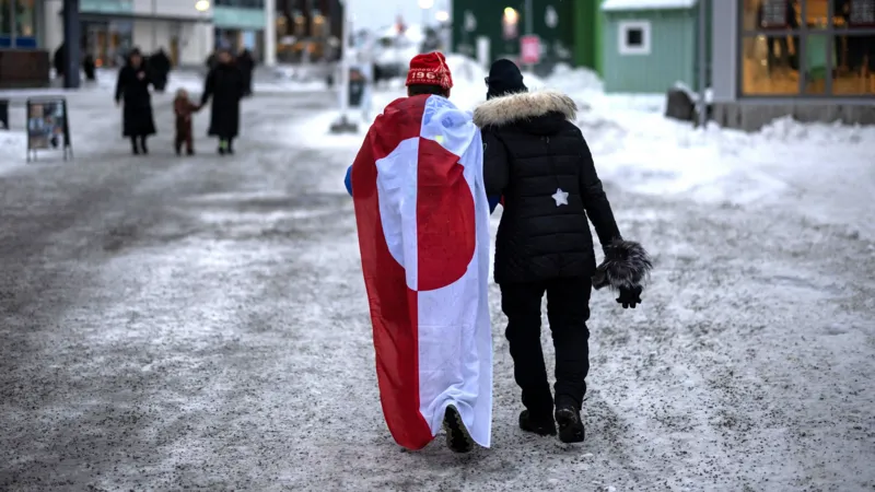 A crowd of protesters holding red and white Greenlandic flags and signs rally at City Square in Copenhagen, Denmark. They are dressed in winter coats, with one man wearing a beanie. A sign at the top of the image reads: "Our land, our freedom, our voice!"