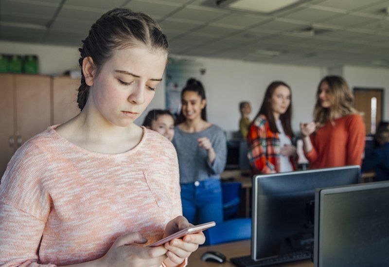 Stock image of a teenage girl looking at a bullying comment on her phone, in a classroom surrounded by other teenagers.