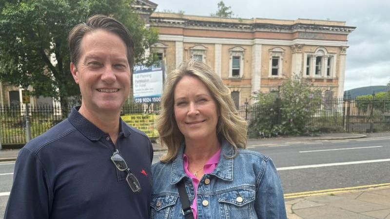 A man, dressed in a navy polo shirt, and a woman wearing a pink top and denim jacket stand in front of the courthouse