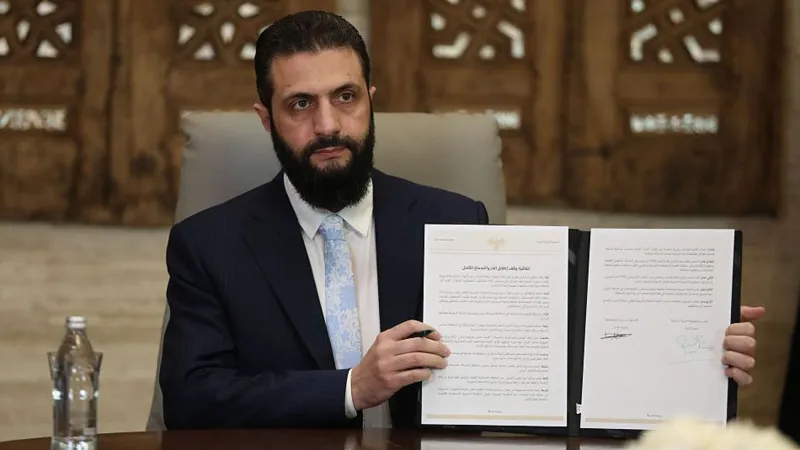 Syrian President Ahmed Sharaa in a navy suit, pale blue tie and white shirt holding up a physical copy of the agreement