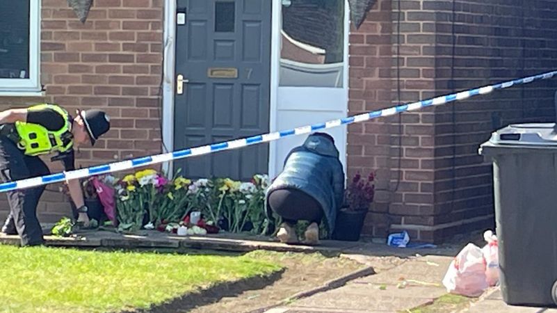 A police officer bends down and another person is knelt down, over a row of flowers laid outside a door. The house is cordoned off by police tape.