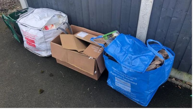 A green food caddy and recycling left out in white and blue bags and a cardboard box