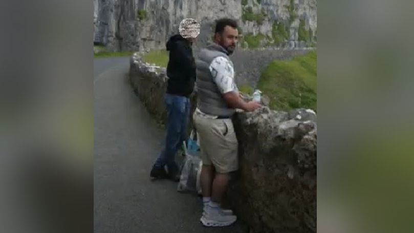 Photo of two men, one close to the camera wearing a grey gillet with beige shorts stood at a rocked wall