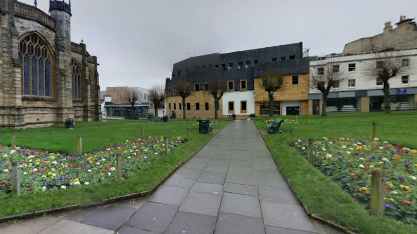 St John's church yard in Yeovil. Buildings can be seen in the background alongside grass and flower beds. There is a path in the middle of the image.