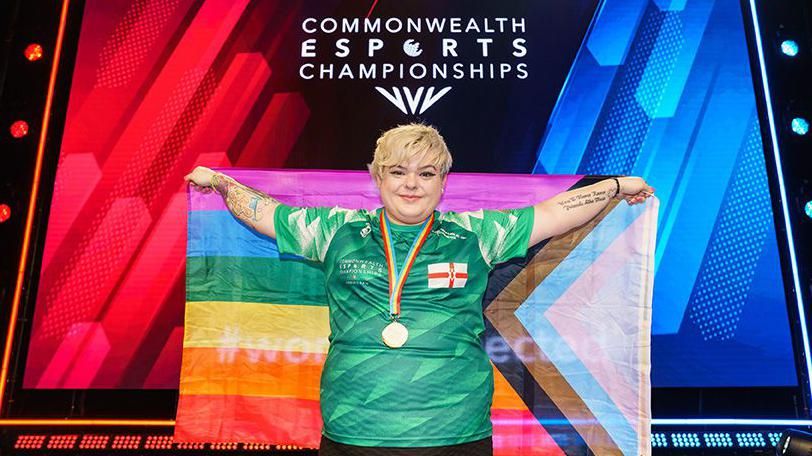 A person in a dark green football shirt with an England St George's cross printed on the right side of the chest holds a rainbow coloured pride flag behind them, arms outstretched. They wear a gold medal around their neck. Behind them a sign reads "Commonwealth esports Championships".