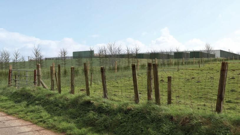 An artist's impression of what the site at West Farm, Aspatria would look like. Green fields surrounded by fencing are foregrounded, with plain boxy buildings in the distance.