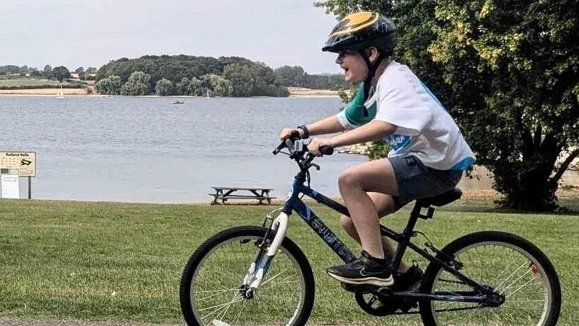 Isaac smiling while riding his bike along a track that runs to the side of a lake. 