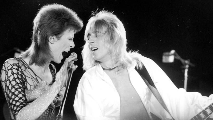Mick Ronson playing guitar and singing into the mic alongside David Bowie
