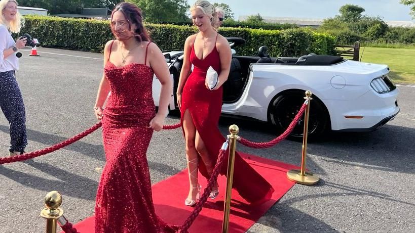 Two pupils walking down the red carpet wearing red dresses rented from the prom shop