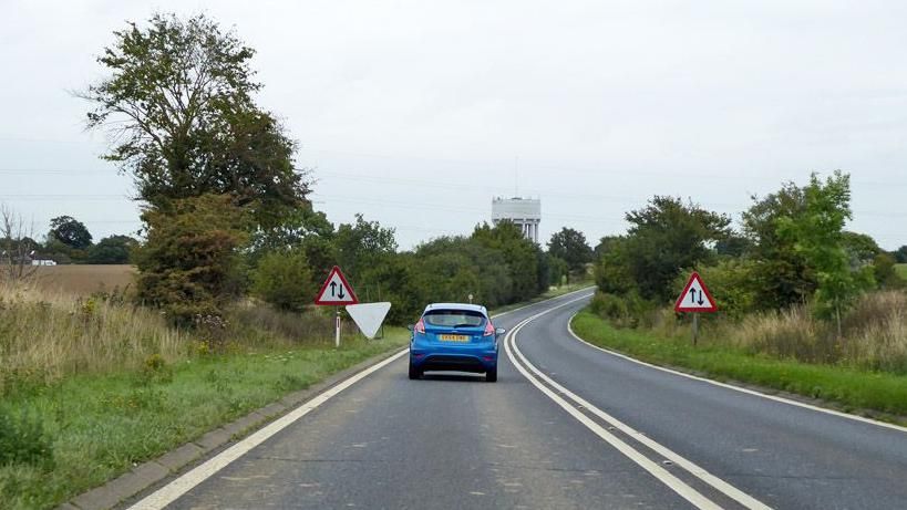 A blue Ford Fiesta driving on the A120, which is a single-carriageway route. Either side of the car are trees and grass.