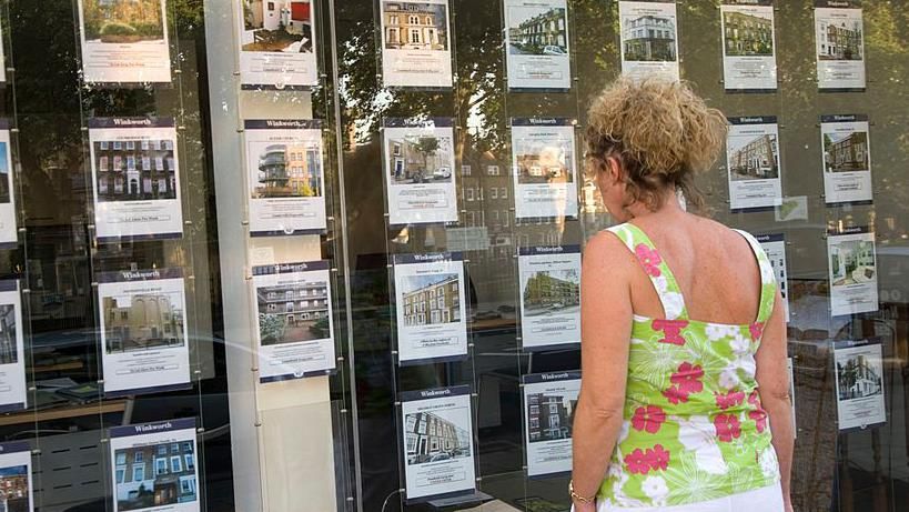 Woman in floral sleeveless top looks in estate agent window at rows of listings