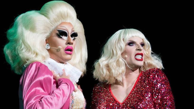 Trixie Mattel performs alongside fellow drag queen Katya. Trixie wears her signature enormous blonde wig and exaggerated make-up with a pink jacket over a white blouse. Katya also wears a blonde wig, paired with red lipstick and a sparkly red dress 