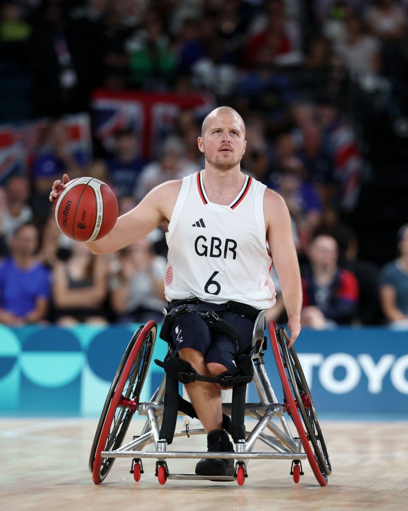 Kyle Marsh wearing a white tank top, sitting in a wheelchair with red and black wheels. He is bouncing a basketball on the court. 