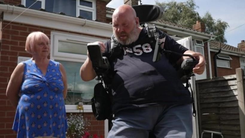 Mick Lloyd pictured in his standing chair with a woman in the background wearing a blue dress. 