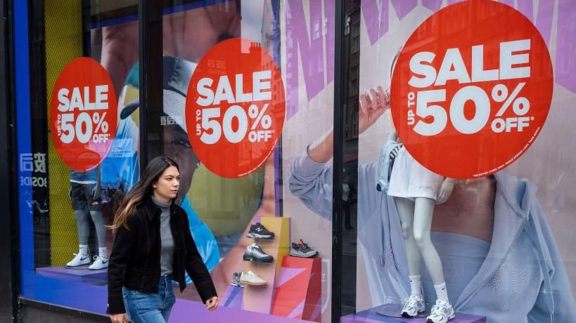 Woman walking past a shop window with '50% off' sale signs