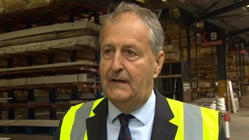 Henry Carver standing in a warehouse, wearing a suit and hi-viz jacket