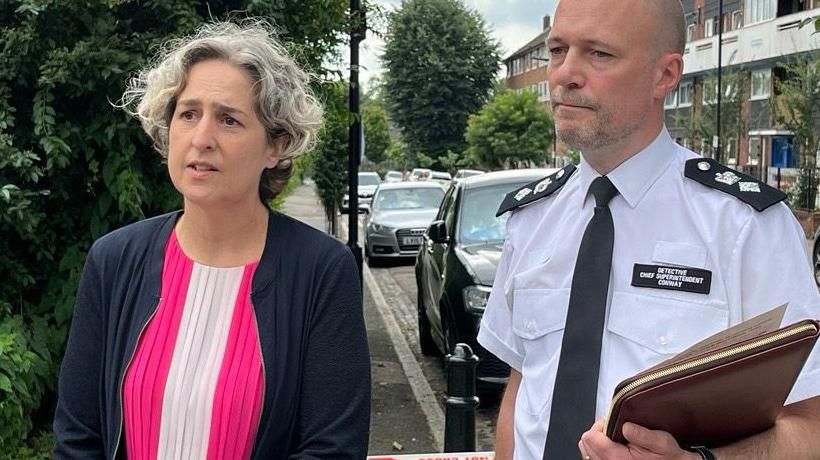 The Mayor of Hackney Caroline Woodley and the Met's Det Ch Insp James Conway stand on a street with parked cars and trees in the background