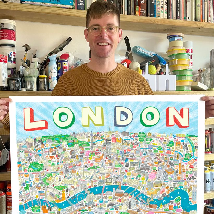 Angry Dan wearing an mustard yellow jumper and glasses standing holding a copy of his illustrated map in his studio