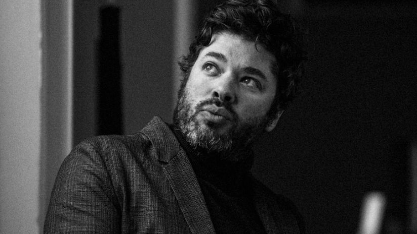 Black and white photo of exhibition creator Darren Emerson, dressed in a jacket and black top. He has dark hair and a beard.