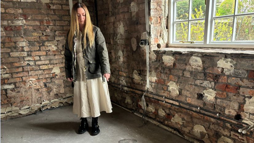 Claire Massey in the kitchen pointing to the spot where the dehumidifier caught fire and melted into the ground. She has long, light hair past her shoulders with a dark jacket on top of a light dress and black shoes. Behind her are brick walls with pipework and electrical wires showing