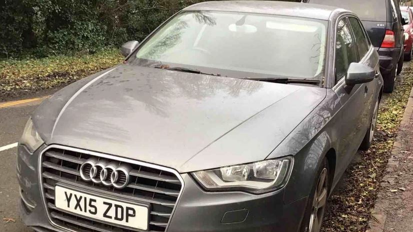 An abandoned grey 2015 Audi A3. It is parallel-parked at the side of a leafy road, with other cars parked up behind it